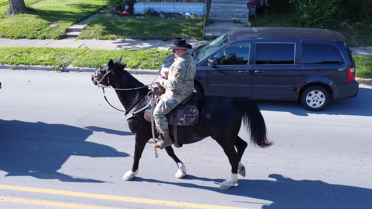 Soldier in camouflage riding a black horse down a neighborhood street.