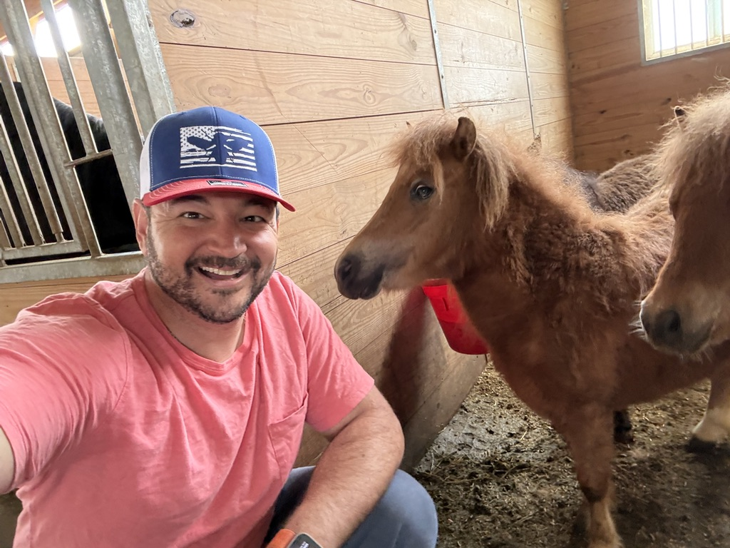 The founder smiles in the barn beside our two miniature ponies.